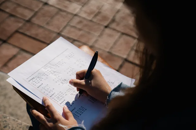 A person writing calculations on a mathematical worksheet with a pen outdoors. Checking the denominator behind support service metrics