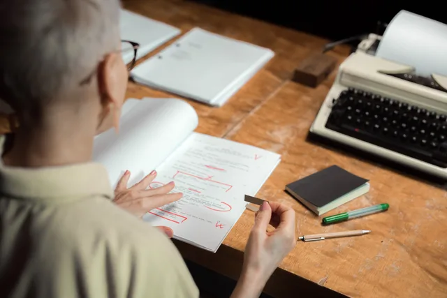 An adult woman reviewing a script with red pen marks at a wooden desk with a typewriter. Reviewing article version and approval controls